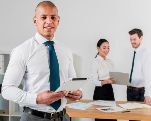 souriant-portrait-jeune-homme-affaires-tenant-tablette-numerique-main-son-collegue-travaillant-arriere-plan_23-2148087266 souriant-portrait-jeune-homme-affaires-tenant-tablette-numerique-main-son-collegue-travaillant-arriere-plan_23-2148087266